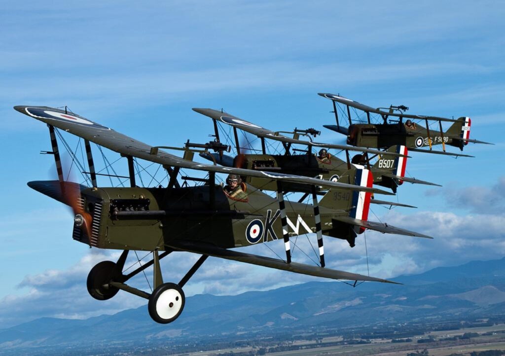 Four SE5a fighters in echelon formation against a blue sky, with mountains and farmland far below.